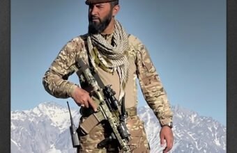 NRF field commander Haseeb Panjshiri standing on a mountain peak in military camouflage gear and a pakol hat holding a rifle against snow-capped Afghan mountains.