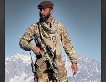 NRF field commander Haseeb Panjshiri standing on a mountain peak in military camouflage gear and a pakol hat holding a rifle against snow-capped Afghan mountains.