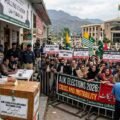 Alt Text An older Kashmiri man with a white beard casts a ballot into a wooden box labeled 'AJK LEGISLATIVE ASSEMBLY ELECTIONS 2026' while a large crowd of protesters with signs demanding 'PUBLIC BOYCOTT' and 'NO REFUGEE SEATS' is separated by police in front of the 'LEGISLATIVES ASSEMBLY' building in Muzaffarabad, Azad Kashmir.