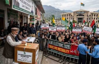 Alt Text An older Kashmiri man with a white beard casts a ballot into a wooden box labeled 'AJK LEGISLATIVE ASSEMBLY ELECTIONS 2026' while a large crowd of protesters with signs demanding 'PUBLIC BOYCOTT' and 'NO REFUGEE SEATS' is separated by police in front of the 'LEGISLATIVES ASSEMBLY' building in Muzaffarabad, Azad Kashmir.