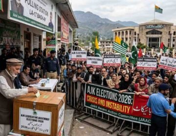 Alt Text An older Kashmiri man with a white beard casts a ballot into a wooden box labeled 'AJK LEGISLATIVE ASSEMBLY ELECTIONS 2026' while a large crowd of protesters with signs demanding 'PUBLIC BOYCOTT' and 'NO REFUGEE SEATS' is separated by police in front of the 'LEGISLATIVES ASSEMBLY' building in Muzaffarabad, Azad Kashmir.