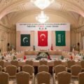 A wide-angle photograph of the prepared venue for the First Meeting of the Joint Committee of the Mecca Defense Agreement in an ornate Istanbul hall, with flags and banners representing Pakistan, Türkiye, and Saudi Arabia.