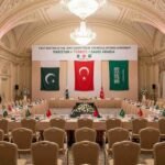 A wide-angle photograph of the prepared venue for the First Meeting of the Joint Committee of the Mecca Defense Agreement in an ornate Istanbul hall, with flags and banners representing Pakistan, Türkiye, and Saudi Arabia.