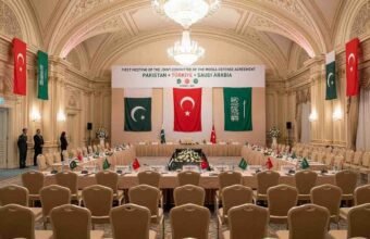 A wide-angle photograph of the prepared venue for the First Meeting of the Joint Committee of the Mecca Defense Agreement in an ornate Istanbul hall, with flags and banners representing Pakistan, Türkiye, and Saudi Arabia.