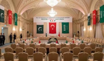 A wide-angle photograph of the prepared venue for the First Meeting of the Joint Committee of the Mecca Defense Agreement in an ornate Istanbul hall, with flags and banners representing Pakistan, Türkiye, and Saudi Arabia.