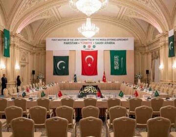 A wide-angle photograph of the prepared venue for the First Meeting of the Joint Committee of the Mecca Defense Agreement in an ornate Istanbul hall, with flags and banners representing Pakistan, Türkiye, and Saudi Arabia.