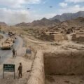 A photojournalistic style image capturing a Pakistani military convoy patrolling a winding road through a dry, mountainous landscape in North Waziristan. The left side features an armored convoy including a leading open-top jeep and several tracked armored personnel carriers (APCs) moving down the paved road. Pakistani soldiers in camouflage uniforms and caps are visible manning the vehicles, with others patrolling the roadside on foot. A prominent green sign at the foreground-left reads "CURFEW IS IN EFFECT, SATURDAY & SUNDAY." On the right, a densely packed village of traditional, deserted mud-brick houses is visible. Two young local children in traditional clothing cautiously peek around the edge of a mud wall in the foreground, watching the passing convoy. In the distance, against the cloudy sky and rugged mountains, two small military drones are visible.