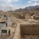 A photojournalistic style image capturing a Pakistani military convoy patrolling a winding road through a dry, mountainous landscape in North Waziristan. The left side features an armored convoy including a leading open-top jeep and several tracked armored personnel carriers (APCs) moving down the paved road. Pakistani soldiers in camouflage uniforms and caps are visible manning the vehicles, with others patrolling the roadside on foot. A prominent green sign at the foreground-left reads "CURFEW IS IN EFFECT, SATURDAY & SUNDAY." On the right, a densely packed village of traditional, deserted mud-brick houses is visible. Two young local children in traditional clothing cautiously peek around the edge of a mud wall in the foreground, watching the passing convoy. In the distance, against the cloudy sky and rugged mountains, two small military drones are visible.