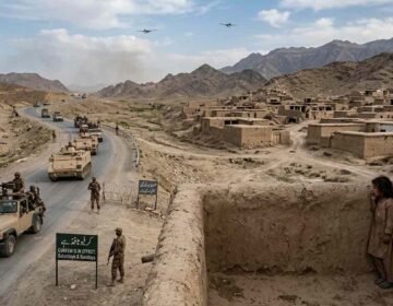 A photojournalistic style image capturing a Pakistani military convoy patrolling a winding road through a dry, mountainous landscape in North Waziristan. The left side features an armored convoy including a leading open-top jeep and several tracked armored personnel carriers (APCs) moving down the paved road. Pakistani soldiers in camouflage uniforms and caps are visible manning the vehicles, with others patrolling the roadside on foot. A prominent green sign at the foreground-left reads "CURFEW IS IN EFFECT, SATURDAY & SUNDAY." On the right, a densely packed village of traditional, deserted mud-brick houses is visible. Two young local children in traditional clothing cautiously peek around the edge of a mud wall in the foreground, watching the passing convoy. In the distance, against the cloudy sky and rugged mountains, two small military drones are visible.