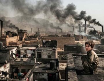 A young child sitting on a dusty rooftop overlooking a semi-urban landscape in Bara and Peshawar, with industrial brick kilns emitting thick black smoke into the hazy sky.