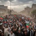 A massive crowd of political demonstrators marching down a dusty street in Pakistan, holding banners and portraits of Imran Khan under an overcast sky.