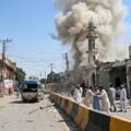 A photograph of a residential street in Pakistan after an explosion. Debris litters the road. A mosque is partially collapsed, with thick smoke rising behind its minaret. A group of men in traditional attire stand distressed on the right, and other people walk on the street.