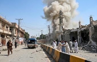 A photograph of a residential street in Pakistan after an explosion. Debris litters the road. A mosque is partially collapsed, with thick smoke rising behind its minaret. A group of men in traditional attire stand distressed on the right, and other people walk on the street.