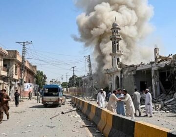 A photograph of a residential street in Pakistan after an explosion. Debris litters the road. A mosque is partially collapsed, with thick smoke rising behind its minaret. A group of men in traditional attire stand distressed on the right, and other people walk on the street.