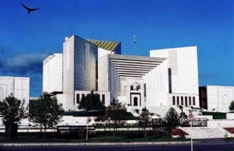 English: Front exterior view of the white marble Supreme Court of Pakistan building in Islamabad under a deep blue sky with a bird flying in the upper-left corner.