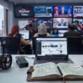 "A detailed shot of a modern newsroom desk in Lahore, Pakistan, with a Canon DSLR camera, Zoom audio recorder, open notebook, and microphone in the foreground. In the background, journalists work at computers facing a large matrix of screens displaying live feeds, stock markets, and the 'Khyber Times' logo. The historic Badshahi Mosque is visible through a large window on the left."