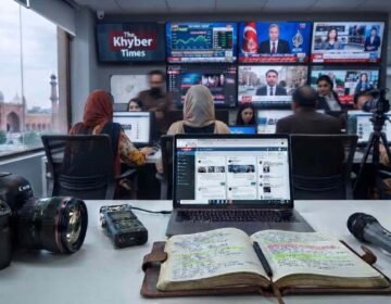 "A detailed shot of a modern newsroom desk in Lahore, Pakistan, with a Canon DSLR camera, Zoom audio recorder, open notebook, and microphone in the foreground. In the background, journalists work at computers facing a large matrix of screens displaying live feeds, stock markets, and the 'Khyber Times' logo. The historic Badshahi Mosque is visible through a large window on the left."