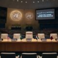 A photograph of the empty United Nations conference hall, prepared for a high-level meeting. The raised stage features a crescent table with five chairs, each with a flag and plaque (USA, Saudi Arabia, PRESIDENT, UAE, Qatar). A large screen on the right displays the text: "POSSIBLE END OF IRAN WAR: POST-WAR STRATEGY DISCUSSION" and Urdu script. Tiers of empty blue seats fill the hall. A timestamp at the bottom right reads: "14:00 EST, Monday, Sep 16, 2026".