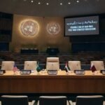 A photograph of the empty United Nations conference hall, prepared for a high-level meeting. The raised stage features a crescent table with five chairs, each with a flag and plaque (USA, Saudi Arabia, PRESIDENT, UAE, Qatar). A large screen on the right displays the text: "POSSIBLE END OF IRAN WAR: POST-WAR STRATEGY DISCUSSION" and Urdu script. Tiers of empty blue seats fill the hall. A timestamp at the bottom right reads: "14:00 EST, Monday, Sep 16, 2026".