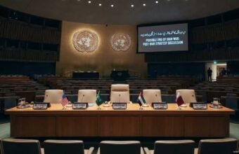 A photograph of the empty United Nations conference hall, prepared for a high-level meeting. The raised stage features a crescent table with five chairs, each with a flag and plaque (USA, Saudi Arabia, PRESIDENT, UAE, Qatar). A large screen on the right displays the text: "POSSIBLE END OF IRAN WAR: POST-WAR STRATEGY DISCUSSION" and Urdu script. Tiers of empty blue seats fill the hall. A timestamp at the bottom right reads: "14:00 EST, Monday, Sep 16, 2026".