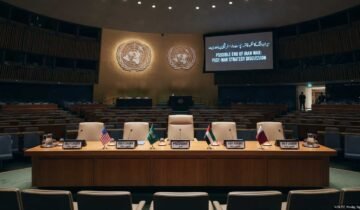 A photograph of the empty United Nations conference hall, prepared for a high-level meeting. The raised stage features a crescent table with five chairs, each with a flag and plaque (USA, Saudi Arabia, PRESIDENT, UAE, Qatar). A large screen on the right displays the text: "POSSIBLE END OF IRAN WAR: POST-WAR STRATEGY DISCUSSION" and Urdu script. Tiers of empty blue seats fill the hall. A timestamp at the bottom right reads: "14:00 EST, Monday, Sep 16, 2026".