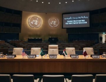 A photograph of the empty United Nations conference hall, prepared for a high-level meeting. The raised stage features a crescent table with five chairs, each with a flag and plaque (USA, Saudi Arabia, PRESIDENT, UAE, Qatar). A large screen on the right displays the text: "POSSIBLE END OF IRAN WAR: POST-WAR STRATEGY DISCUSSION" and Urdu script. Tiers of empty blue seats fill the hall. A timestamp at the bottom right reads: "14:00 EST, Monday, Sep 16, 2026".