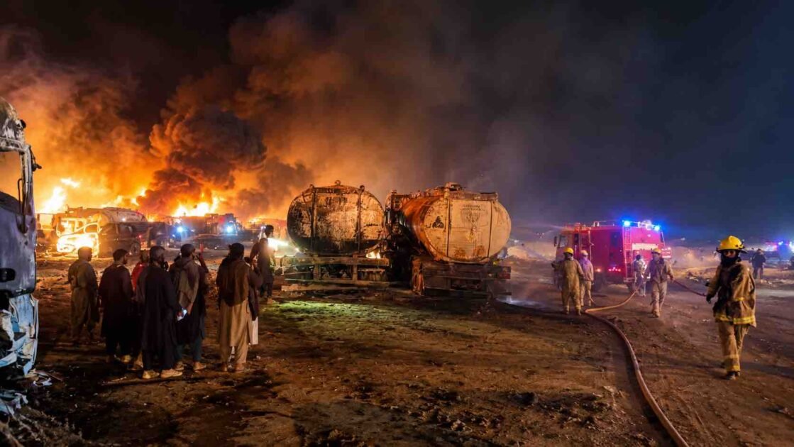 Firefighters battling a massive nighttime fire involving fuel tankers, with thick black smoke, towering flames, and onlookers standing nearby.