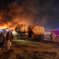 Firefighters battling a massive nighttime fire involving fuel tankers, with thick black smoke, towering flames, and onlookers standing nearby.