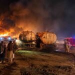Firefighters battling a massive nighttime fire involving fuel tankers, with thick black smoke, towering flames, and onlookers standing nearby.