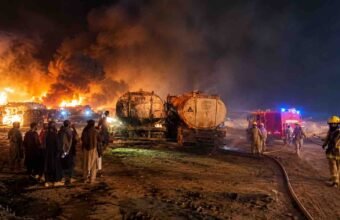 Firefighters battling a massive nighttime fire involving fuel tankers, with thick black smoke, towering flames, and onlookers standing nearby.