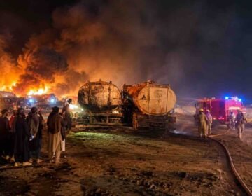 Firefighters battling a massive nighttime fire involving fuel tankers, with thick black smoke, towering flames, and onlookers standing nearby.
