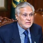Accessibility for visually impaired users and search engines. Describes what is actually visible in the image. Portrait of Pakistani politician Ishaq Dar, with grey hair and beard, wearing a navy suit and checkered shirt, looking intently towards the right while seated in a traditional wooden armchair.