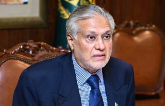 Accessibility for visually impaired users and search engines. Describes what is actually visible in the image. Portrait of Pakistani politician Ishaq Dar, with grey hair and beard, wearing a navy suit and checkered shirt, looking intently towards the right while seated in a traditional wooden armchair.