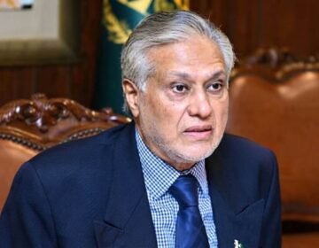 Accessibility for visually impaired users and search engines. Describes what is actually visible in the image. Portrait of Pakistani politician Ishaq Dar, with grey hair and beard, wearing a navy suit and checkered shirt, looking intently towards the right while seated in a traditional wooden armchair.
