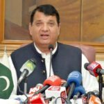 Federal Minister Engineer Amir مقام addressing a press conference in Peshawar, with various news channel microphones and a Pakistani flag on the table