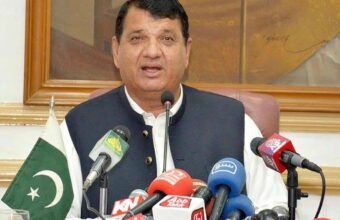 Federal Minister Engineer Amir مقام addressing a press conference in Peshawar, with various news channel microphones and a Pakistani flag on the table