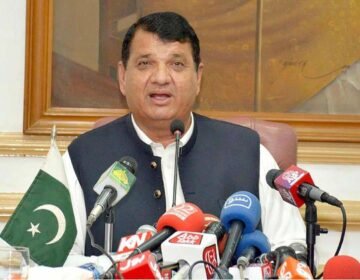 Federal Minister Engineer Amir مقام addressing a press conference in Peshawar, with various news channel microphones and a Pakistani flag on the table