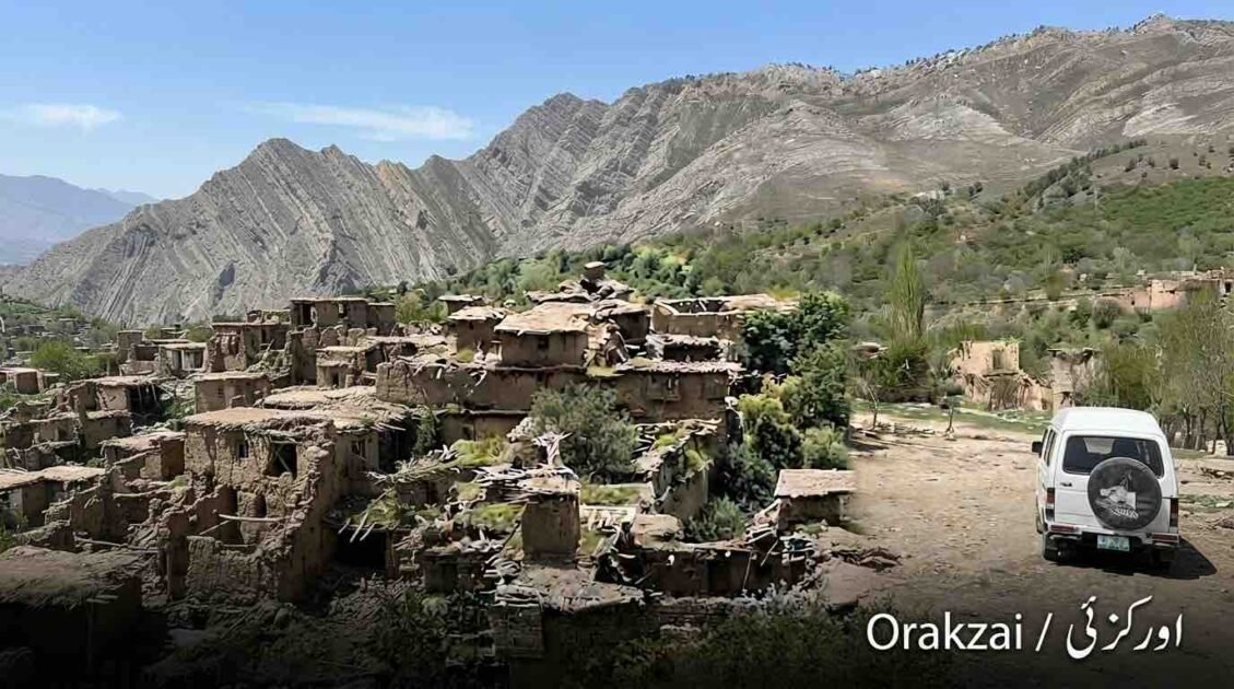 Wide landscape photograph shows an extensive ruined mud-brick village nestled on a green, terraced hillside in the Orakzai district, Pakistan. Towering, rugged, layered mountains dominate the background under a clear blue sky. On the right, a white microvan is parked on a dirt track. The text "Orakzai / اورکزئی" is in the bottom corner.