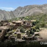 Wide landscape photograph shows an extensive ruined mud-brick village nestled on a green, terraced hillside in the Orakzai district, Pakistan. Towering, rugged, layered mountains dominate the background under a clear blue sky. On the right, a white microvan is parked on a dirt track. The text "Orakzai / اورکزئی" is in the bottom corner.