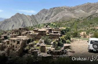 Wide landscape photograph shows an extensive ruined mud-brick village nestled on a green, terraced hillside in the Orakzai district, Pakistan. Towering, rugged, layered mountains dominate the background under a clear blue sky. On the right, a white microvan is parked on a dirt track. The text "Orakzai / اورکزئی" is in the bottom corner.
