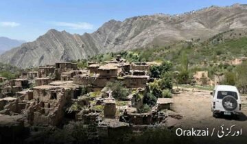 Wide landscape photograph shows an extensive ruined mud-brick village nestled on a green, terraced hillside in the Orakzai district, Pakistan. Towering, rugged, layered mountains dominate the background under a clear blue sky. On the right, a white microvan is parked on a dirt track. The text "Orakzai / اورکزئی" is in the bottom corner.