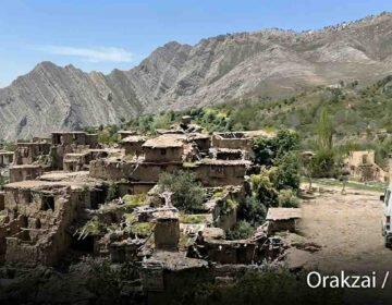 Wide landscape photograph shows an extensive ruined mud-brick village nestled on a green, terraced hillside in the Orakzai district, Pakistan. Towering, rugged, layered mountains dominate the background under a clear blue sky. On the right, a white microvan is parked on a dirt track. The text "Orakzai / اورکزئی" is in the bottom corner.