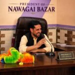 A portrait of a smiling man seated at his office desk with a microphone in front of him. A desk nameplate clearly reads "PRESIDENT NAWAGAI BAZAR". A larger sign on the wall behind him also displays his title: "PRESIDENT OF NAWAGAI BAZAR". Colorful feather dusters are visible on the left side of the desk.