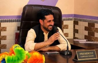 A portrait of a smiling man seated at his office desk with a microphone in front of him. A desk nameplate clearly reads "PRESIDENT NAWAGAI BAZAR". A larger sign on the wall behind him also displays his title: "PRESIDENT OF NAWAGAI BAZAR". Colorful feather dusters are visible on the left side of the desk.
