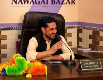 A portrait of a smiling man seated at his office desk with a microphone in front of him. A desk nameplate clearly reads "PRESIDENT NAWAGAI BAZAR". A larger sign on the wall behind him also displays his title: "PRESIDENT OF NAWAGAI BAZAR". Colorful feather dusters are visible on the left side of the desk.
