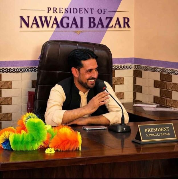 A portrait of a smiling man seated at his office desk with a microphone in front of him. A desk nameplate clearly reads "PRESIDENT NAWAGAI BAZAR". A larger sign on the wall behind him also displays his title: "PRESIDENT OF NAWAGAI BAZAR". Colorful feather dusters are visible on the left side of the desk.