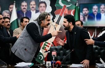 Tensions erupt into a direct confrontation between two men during a Pakistani political meeting, with other attendees attempting to keep them apart amidst a backdrop of political portraits and a fractured party symbol. Alt Text: A phot