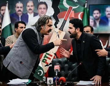 Tensions erupt into a direct confrontation between two men during a Pakistani political meeting, with other attendees attempting to keep them apart amidst a backdrop of political portraits and a fractured party symbol. Alt Text: A phot