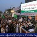 Jamaat-e-Islami Chief Hafiz Naeem ur Rehman addressing a massive public protest rally against petroleum levy with banners and crowd holding party flags in the background.