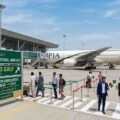 A digital illustration of Islamabad International Airport terminal showing a PIA aircraft on the tarmac, passengers walking, and a green directional sign board detailing the Pak-Afghan transit agreement to the Gulf.